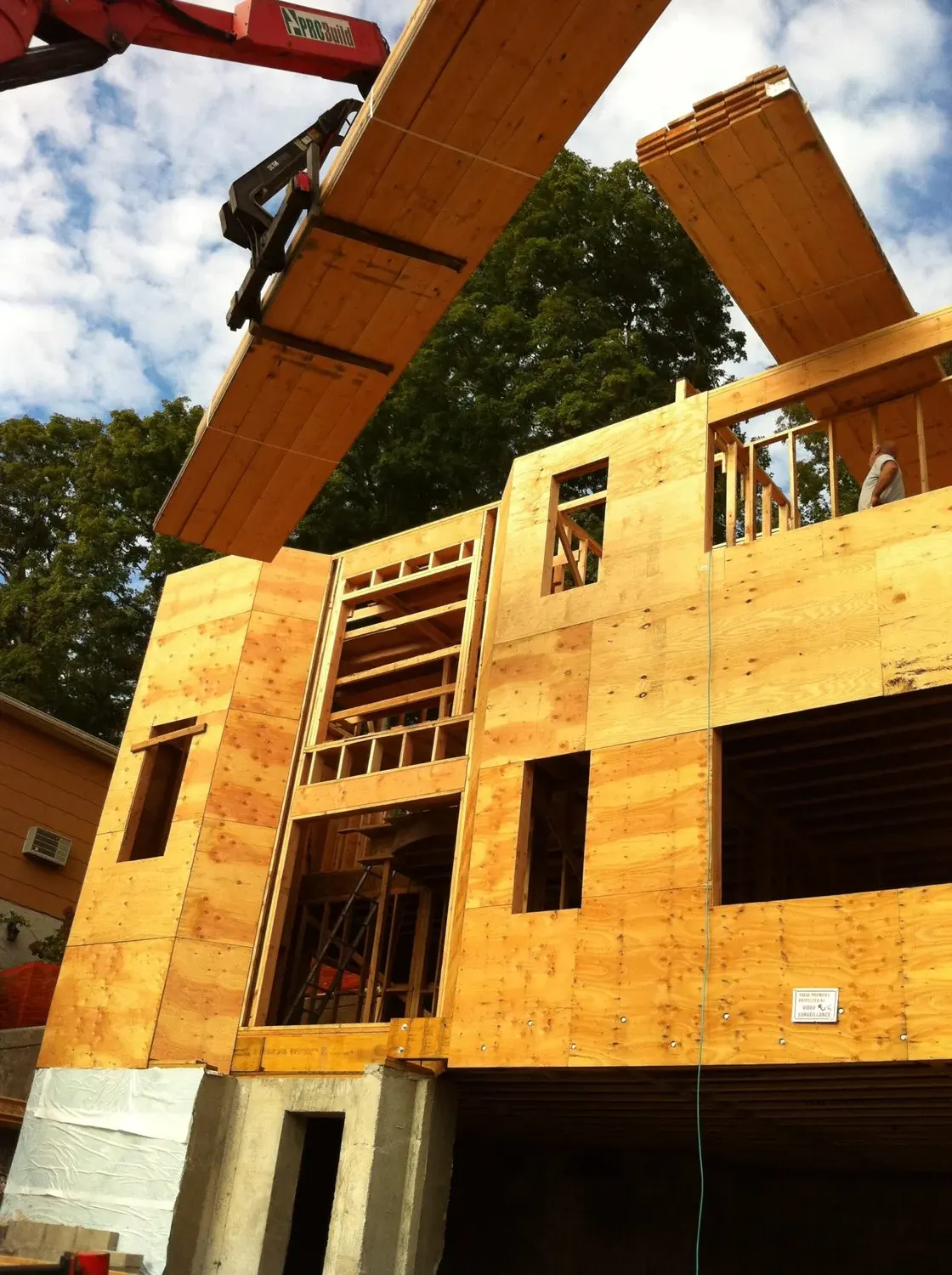 New home construction with exposed framing, plywood sheathing, and overhead crane lifting roof panels
