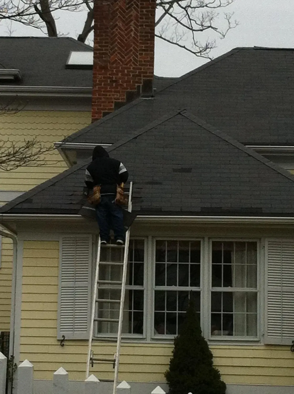 Worker on ladder cleaning or inspecting roof of yellow house with black shingles